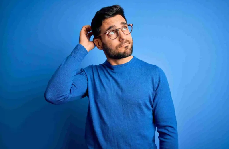 A man with dark hair, a beard, and glasses wearing a blue sweater stands against a blue background, scratching his head and looking upward with a thoughtful expression as if wondering, Can we clear IELTS without coaching?.