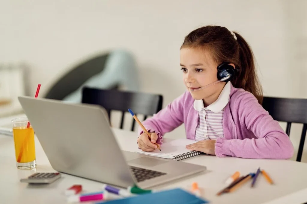 A child in a pink cardigan sits at a desk, using a laptop while taking notes in a notebook, surrounded by colorful stationery and a drink.