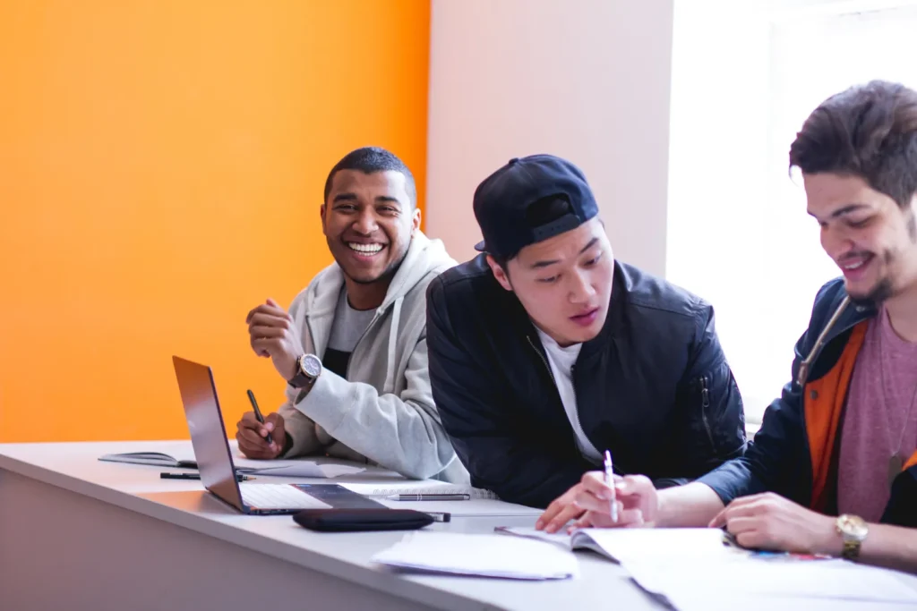 Three young adults collaborate at a table with laptops and notebooks in a brightly lit room featuring an orange wall.