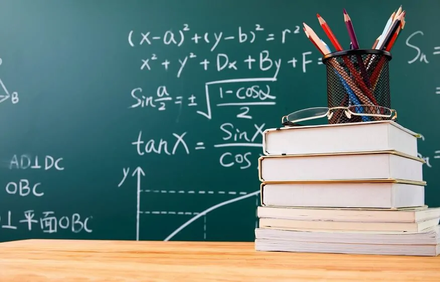 A stack of books with glasses on top, next to a pencil holder, in front of a chalkboard filled with math equations.