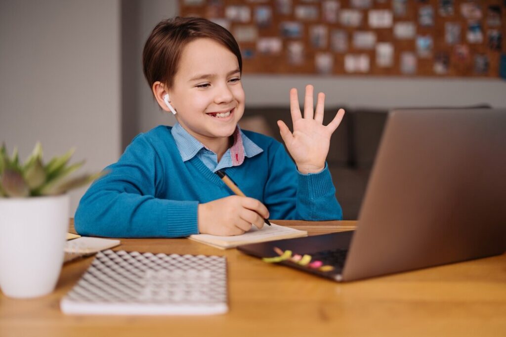 A boy sits at a table with a laptop open and a pen in hand, focused on his work.