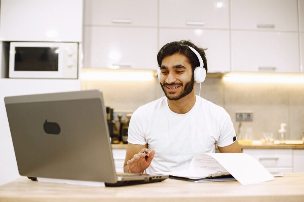 Man in a white t-shirt wearing headphones smiles while video chatting on a laptop in a kitchen. Open notebook and pen in hand suggest study or work.